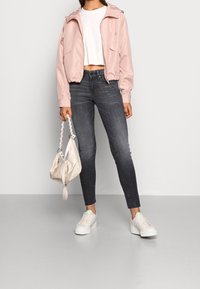 Žena v růžovém krátkém saku, bílém crop topu, tmavě šedých úzkých džínách, bílých teniskách, držící malou bílou kabelku.