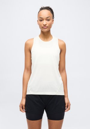 Kobieta z włosami upiętymi w kok, ubrana w białą sportową bluzkę bez rękawów i czarne szorty, stojąca na tle jednolitego jasnego tła.