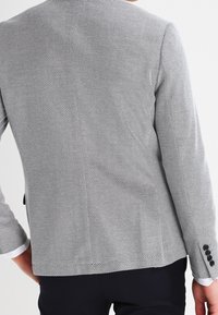 Man som bär en åtsittande svart och vit texturerad kavaj med knäppta manschetter och mörka byxor, visad bakifrån.