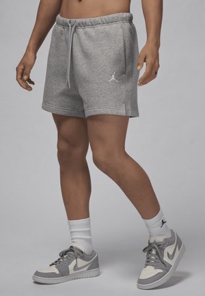 Mężczyzna w szarych sportowych szortach ze sznurkiem, białych skarpetach i szaro-białych butach Nike, stoi na jednolitym tle.