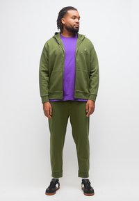 Άνδρας που φοράει κορυφαίο φούτερ με φερμουάρ σε ελιά πράσινο και παντελόνι, με μοβ μπλουζάκι και μαύρα αθλητικά παπούτσια, κοιτάζοντας προς τα πλάγια μπροστά σε λευκό φόντο.