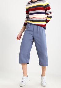 Gestreifter Pullover in Grau, Rot, Gelb und Blau; kombiniert mit hellblauen Cropped-Hosen. Das Model trägt weiße Sneaker.