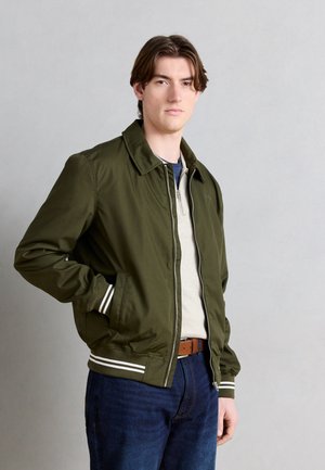 Jeune homme portant une veste bomber vert olive avec des poignets et un col rayés, un pull zippé beige, un jean bleu et une ceinture marron.