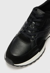 Chaussure de sport noire en cuir texturé et suède avec lacets, dotée d'une semelle intermédiaire blanche et d'un logo de marque discret sur la languette.