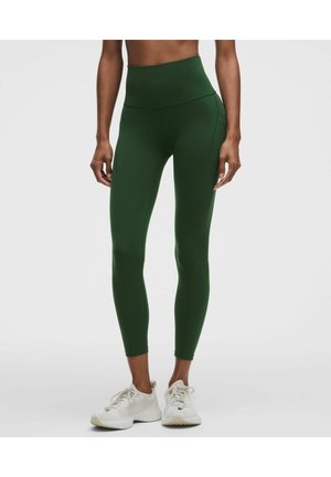 Vrouw draagt donkergroene leggings met hoge taille en witte sportschoenen, staat met ontspannen armen langs haar zij op een effen achtergrond.