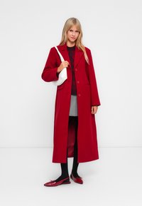 Long red coat with a structured design, two buttons, and side pockets. Worn over a maroon sweater, grey skirt, and black tights with red shoes.