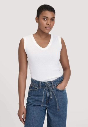 Witte mouwloze V-hals top met contrasterende afwerking, gecombineerd met hooggeknoopte blauwe spijkerbroek met een strikceintuur en zichtbare stikdetails.