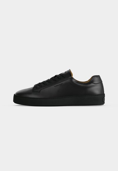 Μαύρο δερμάτινο χαμηλό sneaker με μαύρα κορδόνια, υφασμένο καουτσούκ σόλα και μικρό ανάγλυφο λογότυπο στο πλάι, εμφανίζεται σε προφίλ με γκρι φόντο.