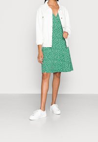 Zielona sukienka w białe kropki, z dekoltem w kształcie litery V, zestawiona z białą dżinsową kurtką i białymi sneakersami. Gładki materiał, luźny krój.