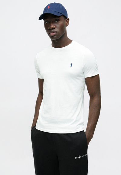 Biała koszulka z krótkim rękawem z małym granatowym haftowanym logo na piersi, w połączeniu z czarnymi spodniami dresowymi ozdobionymi białym brandingiem.