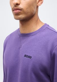 Άνδρας που φοράει μοβ φούτερ με στρογγυλή λαιμόκοψη και την επιγραφή «BORG» κεντημένη στο στήθος, εμφανιζόμενος από τους ώμους μέχρι το στόμα.