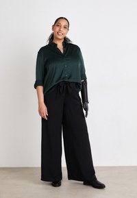 Blouse en satin vert foncé avec détails de boutons, associée à un pantalon large noir. Bras prothétique noir remarquable. Bottines noires sur un fond neutre.