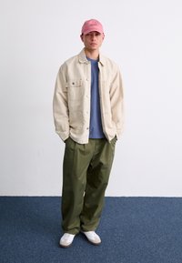 Beige button-up jacket over a blue shirt, paired with green pants and white sneakers. The model wears a pink cap with logo detail.