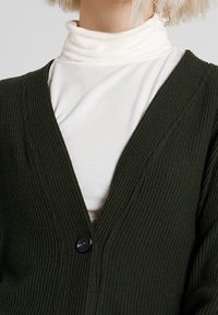 Dunkelgrüner gerippter Cardigan mit tiefem V-Ausschnitt, versehen mit einem einzelnen schwarzen Knopf. Tragen über einem weißen Hochkragenoberteil.