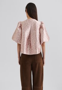 Kurzärmlige Bluse mit Volants aus hellrosa Satin mit einem subtilen Leopardenmuster. Kombiniert mit braunen, hochgeschnittenen, weiten Hosen.