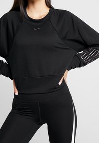 Svart sweatshirt med raglanärmar, prydd med en liten Nike-logotyp och "NIKE"-text i en kontrasterande metallisk finish på ärmen.