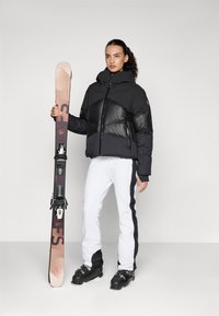 Schwarze Pufferjacke mit glänzenden Kammern, weiße Skihosen mit schwarzen Seitenstreifen und schwarze Skischuhe, hält einen Ski mit Bindungen.