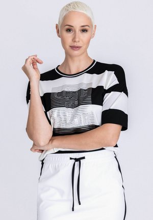 Vrouw met kort blond haar, gekleed in een zwart-wit gestreept shirt met tekst en witte broek met trekkoord, tegen een witte achtergrond.