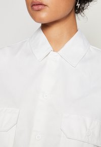 Chemise blanche à boutons avec un col pointu, double poches plaquées, et tissu texturé. Présente des boutons blancs et des lignes épurées.