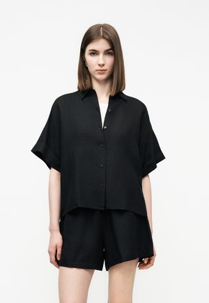 Žena s rovnými hnedými vlasmi po plecia nosí voľnú čiernu košeľu s krátkymi rukávmi na gombíky a zodpovedajúce čierne šortky na bielom pozadí.