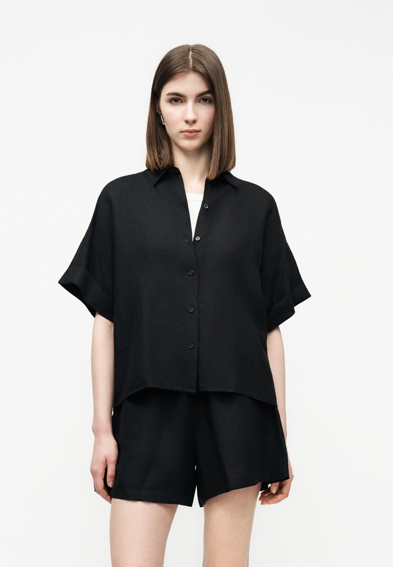 Γυναίκα με ίσια καστανά μαλλιά μέχρι τον ώμο φοράει φαρδιά μαύρη κοντομάνικη μπλούζα με κουμπιά και ταιριαστά μαύρα σορτς σε λευκό φόντο.