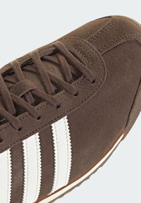 Brun mockasneaker med vita läderstripes, texturerad yta, sydda detaljer och en gummisula med kontrasterande kant.