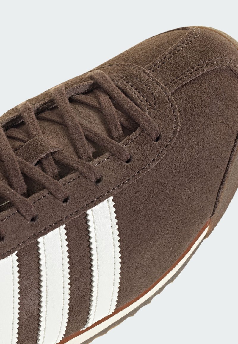 Brun mockasneaker med vita läderstripes, texturerad yta, sydda detaljer och en gummisula med kontrasterande kant.