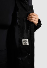 Juodas izoliuotas švarkas su švelnia tekstūra, turintis užtrauktuko kišenę ir etiketę su užrašu: "Šilta apkaba, pagaminta iš audinio."