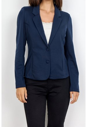 Kvinde iført en tætsiddende marineblå blazer med to knapper over en hvid top med blonder og sorte bukser, stående mod en ensfarvet baggrund.