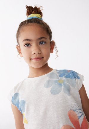 Camisa blanca de manga corta con estampados florales en azul y naranja. Niño con el cabello rizado recogido en un moño, llevando accesorios para el cabello de colores brillantes.