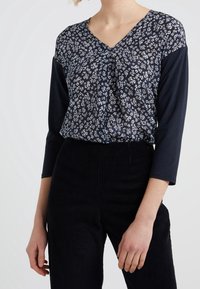 Toppia tummansinistä, jossa on kukkakuosi ja V-pääntie sekä kolmanneksen pituiset hihat, yhdistettynä teksturoituihin mustiin corduroihin housuihin.