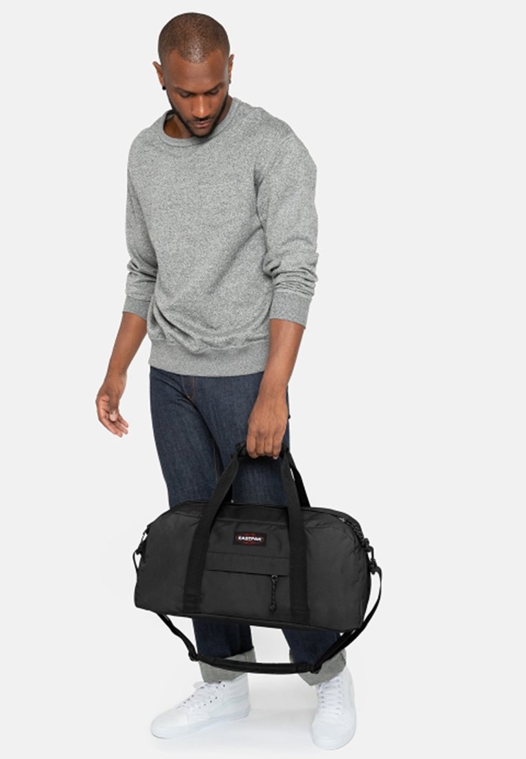 Mustassa duffel-laukussa on kaksi ylhäällä olevaa kantokahvaa, etupuolella vetoketjullinen tasku ja se on valmistettu kestävästä kankaasta. Laukkua kantaa henkilö harmaassa neulepuserossa ja farkuissa.