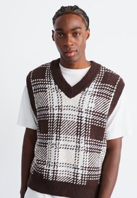 Brown and white checked vest, knit texture, V-neck design, featuring bold patterns, worn over a white T-shirt.