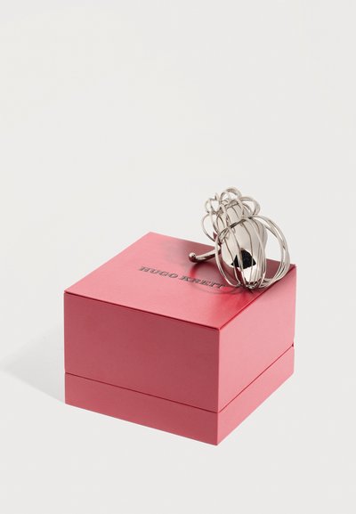 Ασημένιο γλυπτικό δαχτυλίδι με ανοιχτούς βρόχους στην κορυφή ενός κλειστού κόκκινου κουτιού Hugo Kreit, εμφανίζεται σε λευκό φόντο.