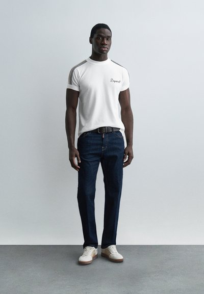 Άνδρας που φοράει λευκό μπλουζάκι με σχέδιο στους ώμους, σκουρόχρωμο μπλε τζιν, μαύρη ζώνη και λευκά αθλητικά παπούτσια, στέκεται μπροστά σε απλό γκρι φόντο.