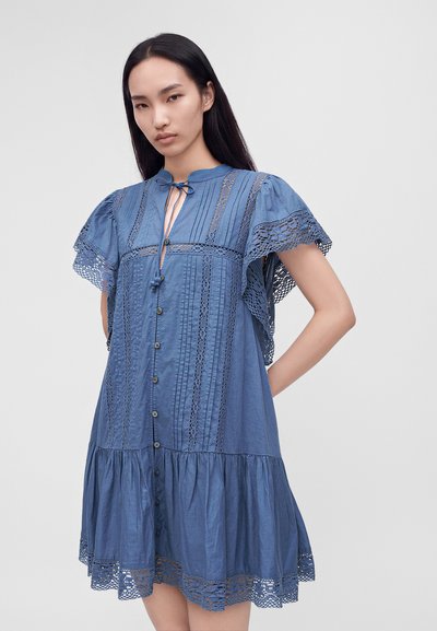 Femme portant une robe bleue boutonnée avec des détails en dentelle sur les manches et l'ourlet, debout avec une main sur la hanche devant un fond uni.