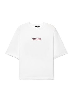 T-shirt blanc à manches courtes avec un texte bordeaux "CHAOS LOOKS GOOD ON ME" centré sur la poitrine et une petite étiquette noire à l'intérieur du col.