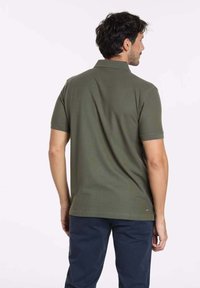 Polo-shirt vert olive à manches courtes, col classique et coutures arrière. Fabriqué en coton. Porté avec un pantalon bleu marine.