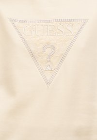 Υφασμα σε κρέμ απόχρωση με ανάγλυφο τριγωνικό λογότυπο. Διακοσμημένο με στρας και σχέδιο δαντέλας μέσα στο τρίγωνο. Περιλαμβάνει τα κείμενα «GUESS» και «U.S.A.»