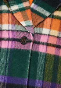 Nærbillede af en uldne ternet frakke med orange, grønne, pink, lilla og hvide tern samt en enkelt mørkebrun knap fastgjort nær kraven.