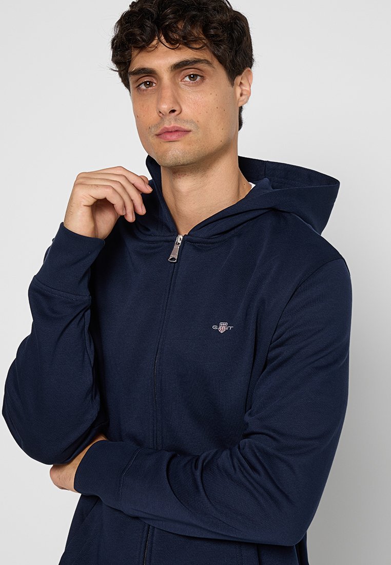 Marinblå zip-up hoodie i mjukt tyg, med huva, långa ärmar och en liten logotyp på bröstet. Slät textur med en figurnära design.