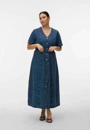 Frau, die den Bindegürtel eines blauen, knopf-geschlossenen Midi-Jeanskleids mit kurzen Ärmeln und V-Ausschnitt richtet, trägt schwarze Riemchensandalen.