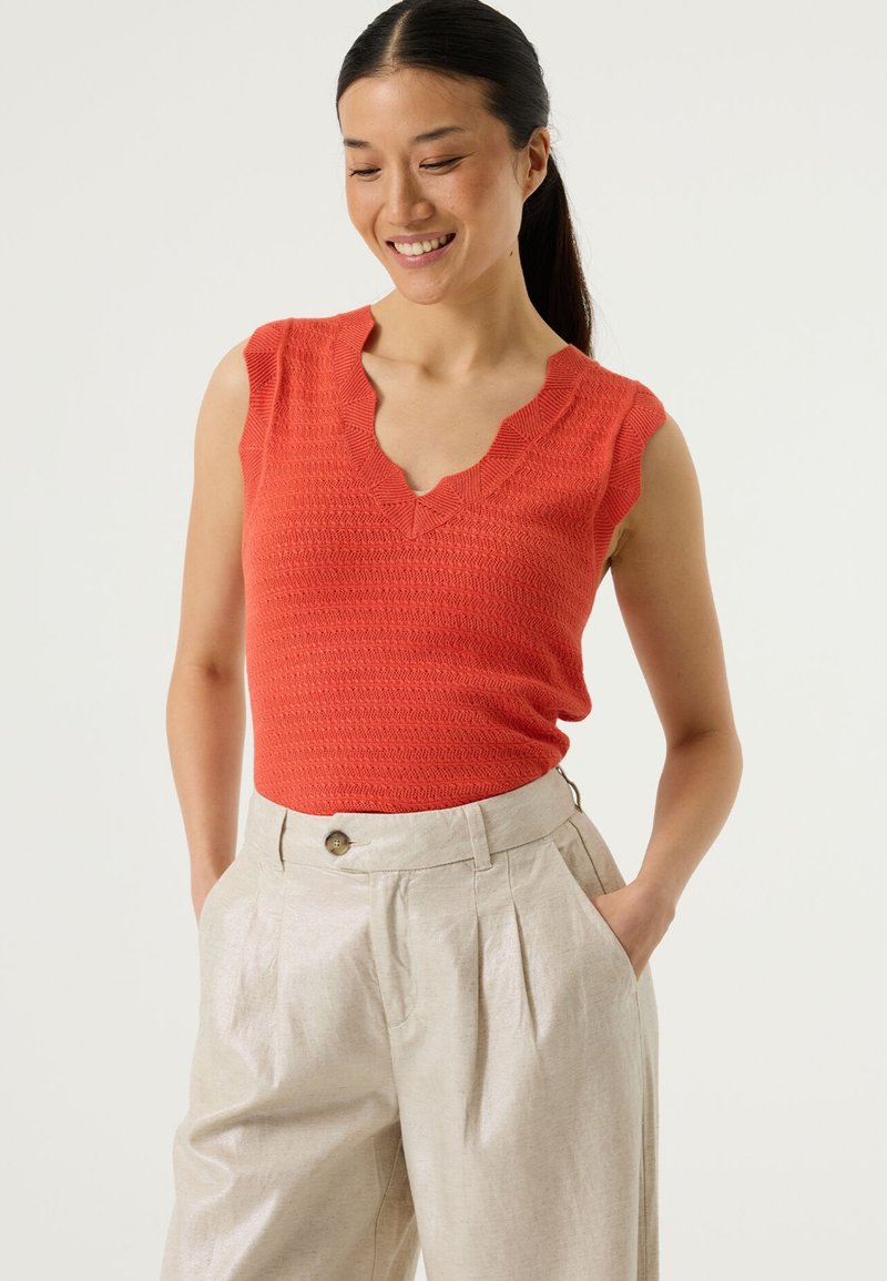 Haut rouge sans manches avec un décolleté festonné et un motif texturé, porté avec un pantalon taille haute beige clair avec un détail de bouton.