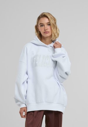 Bílá oversized mikina vyrobená z měkké tkaniny, s předním grafickým motivem posetým rhinestones a klokaní kapsou. Ribbed manžety a lem.