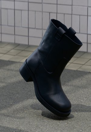 Stivaletto alla caviglia in pelle nera con tacco basso, posato su un terreno texturizzato vicino a una parete piastrellata sullo sfondo.