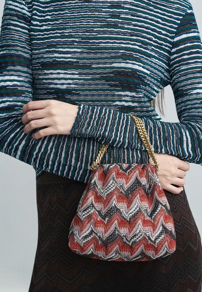 Person wearing a blue-green striped sweater and brown skirt, holding a red, black, and white zigzag patterned bag with a gold chain strap.