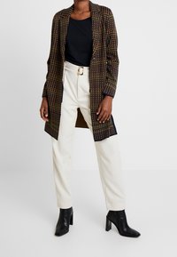 Manteau long en tartan marron avec des accents multicolores, porté sur un haut noir et un pantalon blanc, accompagné de bottines noires.