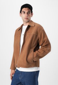 Brun mockaläder zip-jacka med en krage, sidofickor och avslappnad passform. Bärs över en vit t-shirt och blå jeans.