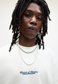 White sweatshirt with blue embroidered text. Two layered necklaces: one pearl and one metallic silver. Dark, twisted hair and minimal facial features.