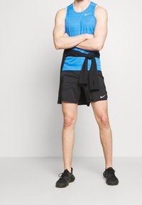 Herre activewear sæt: blå stribet tanktop, sorte shorts med Nike-logo, sort sweater bundet om taljen og sorte sportsko.